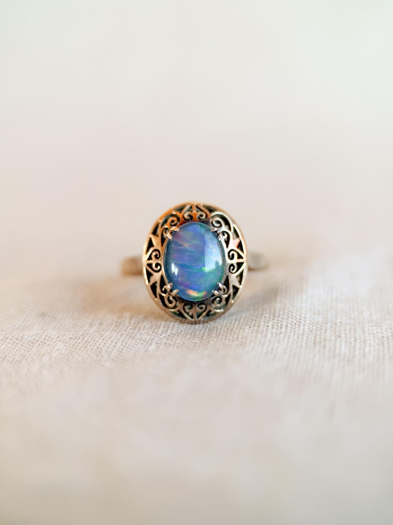 May include: A silver ring with an oval, iridescent blue gemstone. The gemstone is set in a decorative, filigree-style bezel. The ring is displayed on a white, textured surface, showcasing the jewelry's design and color.