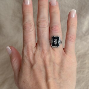 May include: A silver ring with a black center stone and small white accent stones. The ring is on a hand with manicured nails.