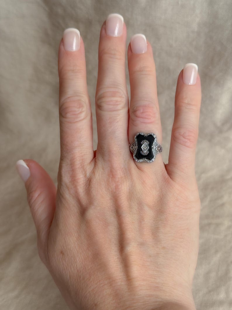 May include: A silver ring with a black center stone and small white accent stones. The ring is on a hand with manicured nails.