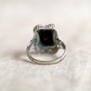 May include: A silver ring with an ornate design. The ring features a dark, square-shaped stone at the center, surrounded by smaller, clear stones. The band has delicate etched details. The ring is set against a soft, neutral background.
