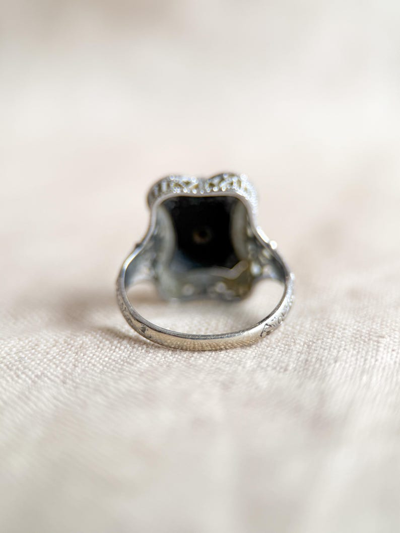 May include: A silver ring with an ornate design. The ring features a dark, square-shaped stone at the center, surrounded by smaller, clear stones. The band has delicate etched details. The ring is set against a soft, neutral background.