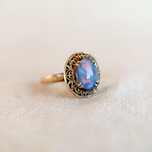 May include: A gold ring featuring an oval, iridescent blue opal gemstone. The opal is set within a decorative, filigree-style bezel. The ring is displayed on a neutral-colored, textured surface, highlighting the jewelry's details.
