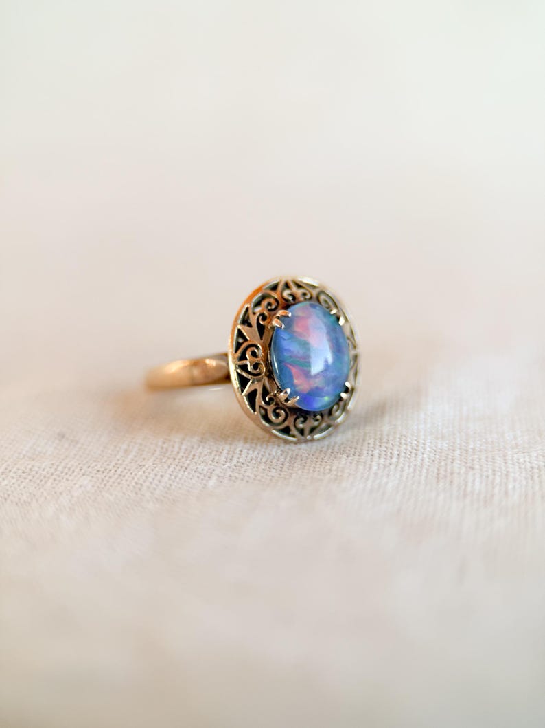 May include: A gold ring featuring an oval, iridescent blue opal gemstone. The opal is set within a decorative, filigree-style bezel. The ring is displayed on a neutral-colored, textured surface, highlighting the jewelry's details.