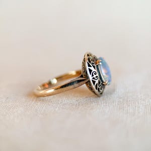 May include: A gold ring with an oval, iridescent gemstone. The gemstone is set in a silver filigree bezel. The ring band is a simple, polished gold. The ring is set against a neutral background.
