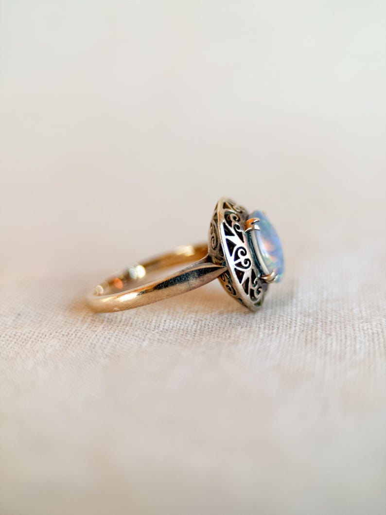 May include: A gold ring with an oval, iridescent gemstone. The gemstone is set in a silver filigree bezel. The ring band is a simple, polished gold. The ring is set against a neutral background.