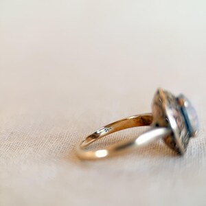 May include: A gold ring with an ornate, square-shaped setting and a light-colored stone. The ring band is thin and smooth, with the marking "18k" visible. The ring is set against a neutral background.