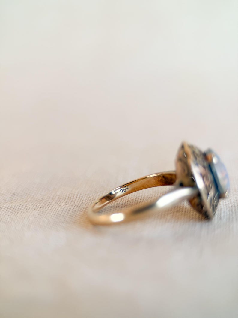 May include: A gold ring with an ornate, square-shaped setting and a light-colored stone. The ring band is thin and smooth, with the marking "18k" visible. The ring is set against a neutral background.