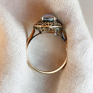 May include: A vintage-style ring with a light blue oval gemstone set in an ornate, filigree-detailed setting. The ring has a gold band and a detailed design around the gemstone, showcasing a classic jewelry aesthetic.