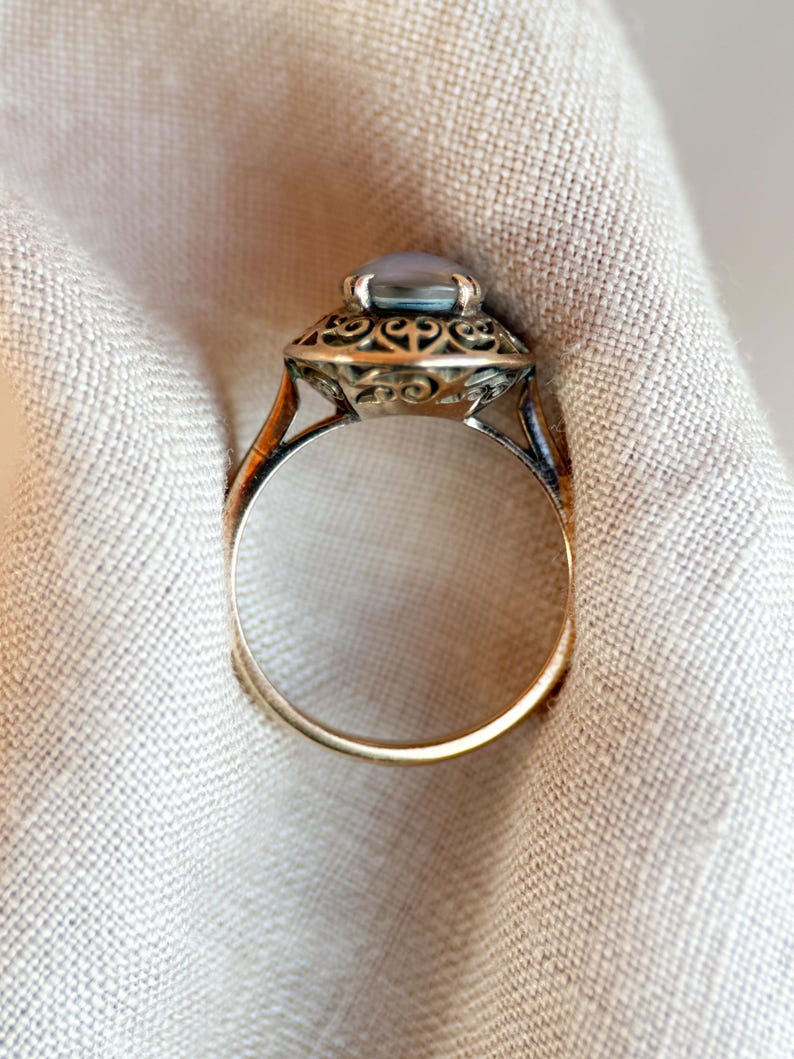 May include: A vintage-style ring with a light blue oval gemstone set in an ornate, filigree-detailed setting. The ring has a gold band and a detailed design around the gemstone, showcasing a classic jewelry aesthetic.