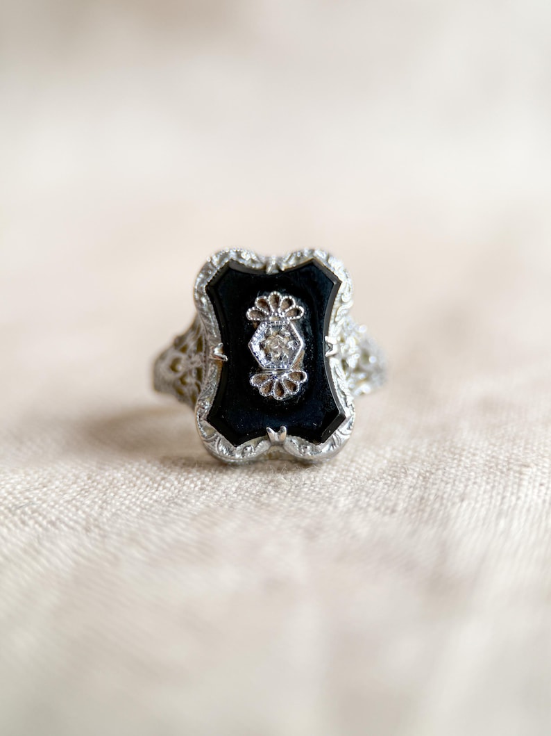 May include: An antique silver ring with a rectangular black onyx center stone. The onyx is framed by a detailed silver filigree design. A small diamond is set in the center of the onyx, adding a touch of sparkle. The ring has an Art Deco style.