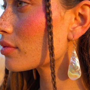 May include: A teardrop-shaped earring featuring an iridescent, pearlescent shell surface with a smooth, polished finish. The pendant hangs from a thin, gold-toned metal hook that passes through the earlobe, reflecting light with soft rainbow hues.