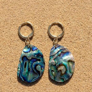 May include: Two gold-coloured hoop earrings, each with a teardrop-shaped pendant. The pendants are crafted from iridescent abalone shell, showcasing blue, green, and purple hues. The earrings are displayed on a sandy surface.
