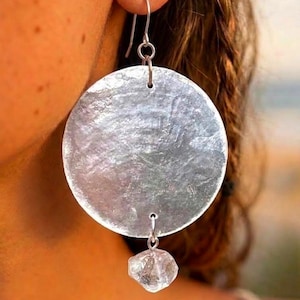 May include: A silver-toned earring featuring a large, circular, hammered metal disc suspended from a simple hook. A small, clear, irregularly shaped, faceted stone charm hangs from the bottom center of the disc, creating a dangling design.