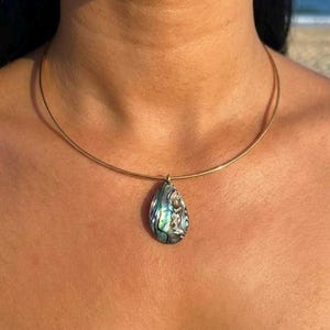 May include: A gold-toned metal collar necklace featuring a teardrop-shaped pendant made of iridescent abalone shell. The pendant displays a swirling pattern of green, blue, and purple hues with a polished finish, suspended from a thin, rigid wire band.