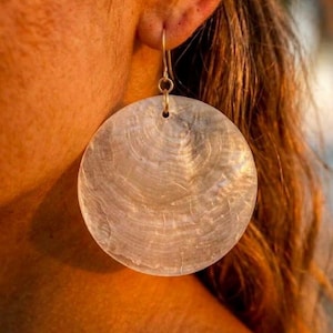 May include: A large, circular earring made of iridescent, translucent shell material. The disc features a natural, concentric growth pattern with a smooth, polished surface. It hangs from a simple, thin metal hook that passes through the earlobe.