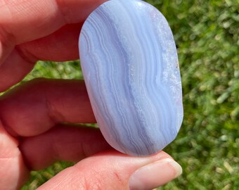 May include: A polished, oval-shaped blue lace agate stone. The stone features bands of light blue and white, creating a layered pattern. The smooth surface reflects light, highlighting the natural variations in color and texture. The stone is held in a hand.