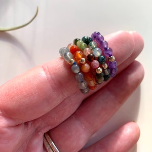 May include: A stack of colourful gemstone rings with gold beads. The rings are made of amethyst, green aventurine, carnelian, sunstone, labradorite, and a dark grey stone.