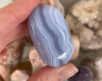 May include: A smooth, oval-shaped blue lace agate gemstone. The stone features bands of light blue and white, creating a layered pattern. The polished surface reflects light, highlighting the natural variations in the stone.