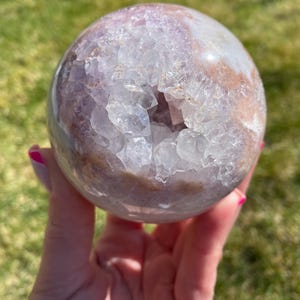 May include: A spherical crystal geode with a polished exterior. The geode features a mix of translucent and opaque sections, with a cavity filled with sparkling crystal formations. The colours range from pale lavender to light brown.
