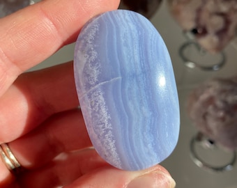 May include: A smooth, oval-shaped blue lace agate stone with light blue and white banding. The polished gemstone is held in a hand, showcasing its natural patterns and colors. Other stones are visible in the background.