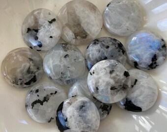 May include: A collection of ten round, white moonstone cabochons with black inclusions. The stones are smooth and polished, and they have a slightly iridescent sheen.