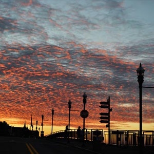 May include: Silhouette of a bridge with streetlights against a vibrant orange and pink sunset sky. The sky is filled with fluffy clouds.