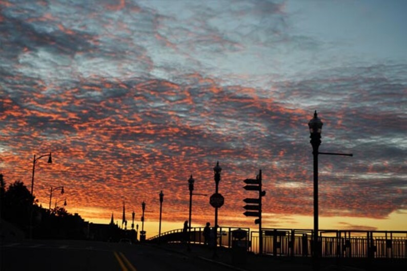 May include: Silhouette of a bridge with streetlights against a vibrant orange and pink sunset sky. The sky is filled with fluffy clouds.