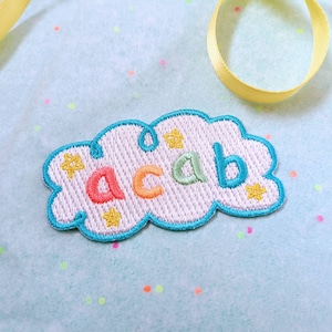 Puede incluir: Un parche bordado de color azul claro con forma de nube con la palabra "acab" en colores arcoíris. El parche tiene estrellas amarillas alrededor de los bordes.