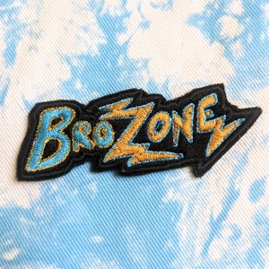 May include: A black embroidered patch with the text "Bro Zone" in gold and blue lettering. The text is stylized with a lightning bolt design.