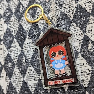 May include: A clear plastic keychain featuring a cartoon girl with red hair and a blue and white dress. The girl is inside a small wooden house with a sign that reads "Do Not Shake".