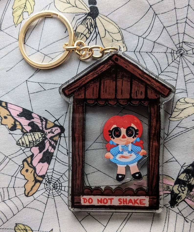 May include: A red-haired doll with X's for eyes is trapped inside a wooden frame with the words "Do Not Shake" printed on the bottom. The doll is wearing a blue and white gingham dress with a red bow and a white apron with brown stains. The frame is attached to a gold key chain.