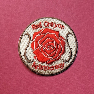 Może przedstawiać: Naszywka haftowana w kolorze beżowym z czerwoną różą i tekstem "Red Crayon Aristocracy".