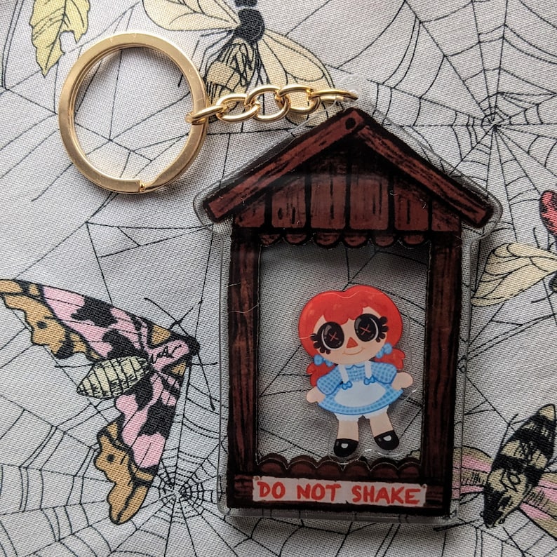May include: A red-haired doll with a blue and white dress is inside a clear plastic frame with a brown wooden frame. The frame has a gold chain attached to it and the text "Do Not Shake" is printed on the bottom.