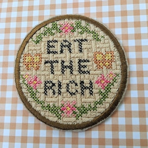 May include: A beige round embroidered patch with a brown border. The patch features a floral design with green leaves and pink and orange flowers. The text "Eat the Rich" is embroidered in black.