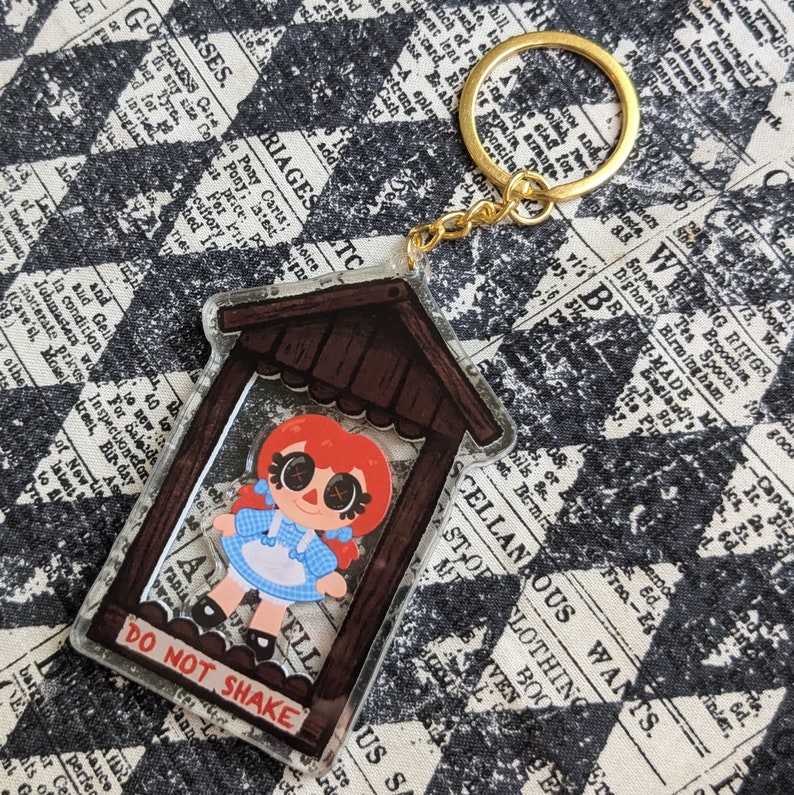 May include: A clear acrylic keychain featuring a cartoon illustration of a red-haired girl in a blue and white gingham dress. The girl is inside a small wooden structure with the text "DO NOT SHAKE" on the bottom. The keychain is attached to a gold metal key ring.