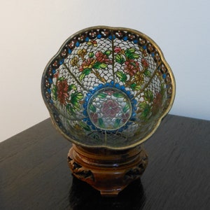 Puede incluir: Un cuenco decorativo con un diseño floral, colocado sobre un soporte de madera tallada. El cuenco presenta un delicado patrón calado con motivos florales rojos, rosas, amarillos y verdes. El borde está adornado con oro.