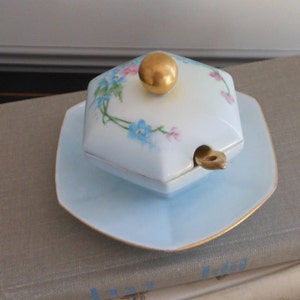 May include: A light blue porcelain sugar bowl with a matching saucer. The hexagonal lid features a gold knob and is decorated with blue and pink floral accents. The saucer and lid are trimmed with gold.