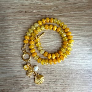 May include: A layered necklace featuring yellow and gold beads. The necklace has a gold clasp and a charm cluster with seashells and a pearl. The necklace is displayed on a wooden surface.