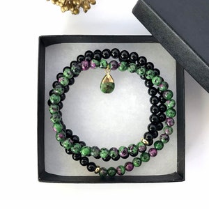 May include: A black box with a green and pink gemstone bracelet and a gold pendant inside. The bracelet is made of small round beads.