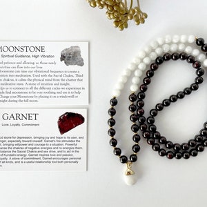 May include: A mala or prayer bead necklace with white moonstone and garnet beads. The necklace is on a white surface. The card on the left describes the properties of moonstone and garnet crystals. "Moonstone: Intuition, Spiritual Guidance, High Vibration" and "Garnet: Love, Loyalty, Commitment" are written on the cards.