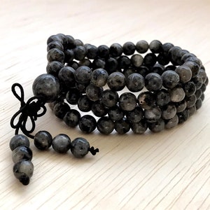 Puede incluir: Un brazalete negro de cuentas con un borla anudada. Las cuentas están hechas de labradorita, una piedra preciosa conocida por sus propiedades iridiscentes.