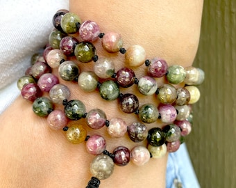 May include: A multi-strand bracelet made of round, multi-colored gemstone beads. The beads are a mix of pink, green, and brown with a speckled pattern. The bracelet is tied with a black cord.