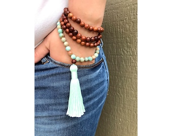 May include: A layered mala necklace with brown wooden beads and turquoise-colored beads. The necklace has a light green tassel.