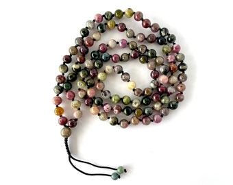 May include: A strand of colorful tourmaline beads in shades of green, pink, brown, and yellow. The beads are strung on a black cord, forming a necklace or prayer beads. The beads are round and smooth, with a natural, earthy appearance.