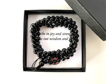 Può includere: Una collana di mala di onice nero in una scatola regalo nera. La scatola contiene un biglietto bianco con il testo "the in joy and strength, the out wisdom and peace".