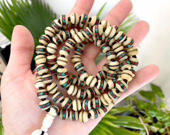 May include: Cream-colored beaded necklace with red and turquoise accents. The beads are cylindrical with decorative bands of color. The necklace is held in a hand, with a white tassel at the end. The beads are strung on a black cord.