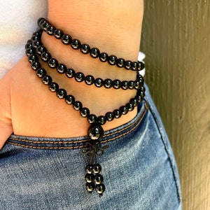 May include: A black beaded bracelet with a decorative knot and tassel detail. The bracelet is made of small, round, glossy black beads and is wrapped around a wrist. The bracelet is worn with a white shirt and blue jeans.