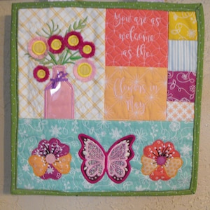May include: A quilted wall hanging with a green border and a patchwork design. The center features a quote: "You are as welcome as the flowers in May." The design includes embroidered flowers, a butterfly, and a vase.