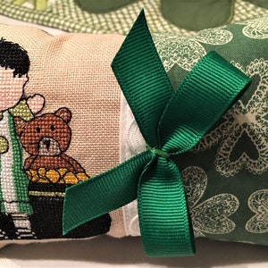 May include: A small pillow with a green and white striped design featuring a boy holding a teddy bear. The pillow is tied with a green ribbon and has a green shamrock pattern on the back.