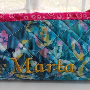 May include: A teal blue quilted zippered pouch with a pink and white polka dot trim. The pouch has a gold embroidered name "Marta" on the front.